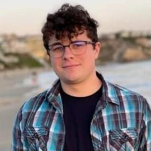 A pale man with brown hair and glasses wearing a blue flannel and a black shirt smiles in front of a beach
